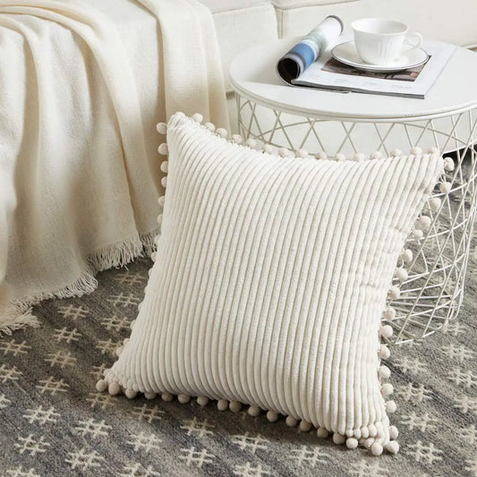 Beige pillow with pom-pom trim on a couch with a side table in the background