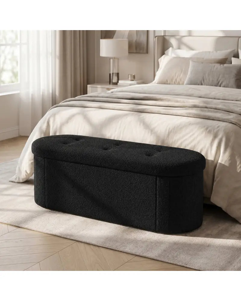 A black upholstered storage bench with a tufted lid sits at the foot of a neatly made bed in a softly lit bedroom.