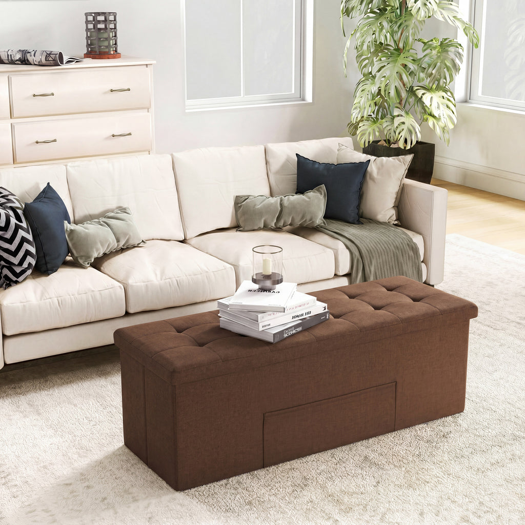 A brown tufted fabric storage ottoman bench with built-in drawers, styled in a bright living room on a white rug in front of a cream-colored sectional sofa.