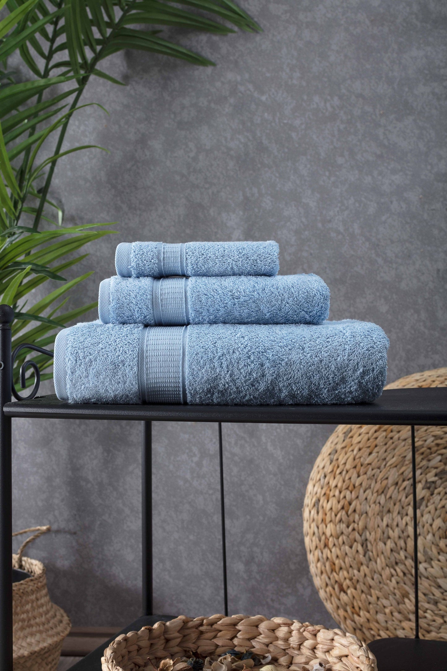 Stack of blue towels on a black shelf against a gray wall with plants and woven baskets.