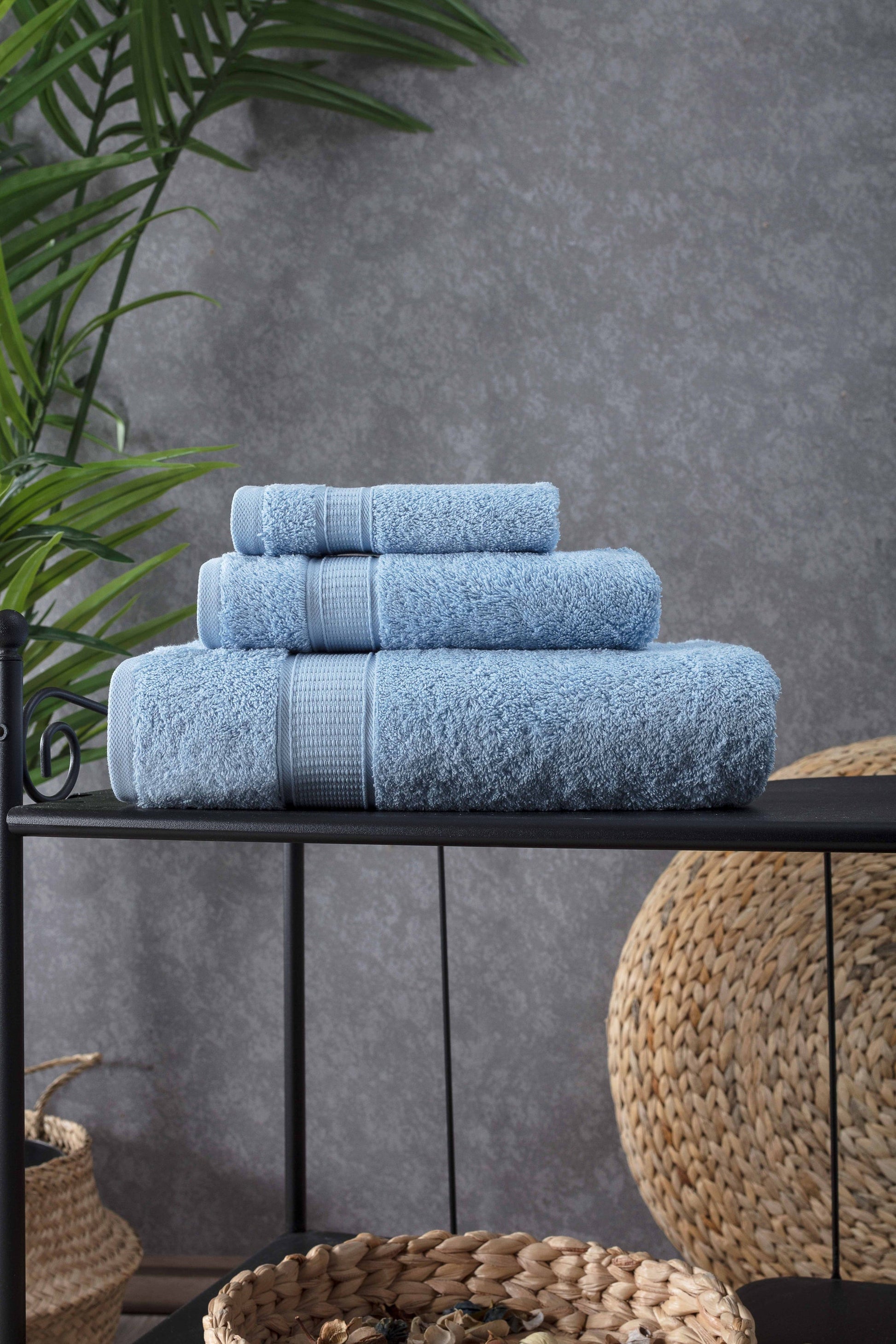 Stack of blue towels on a black shelf against a gray wall with plants and woven baskets.