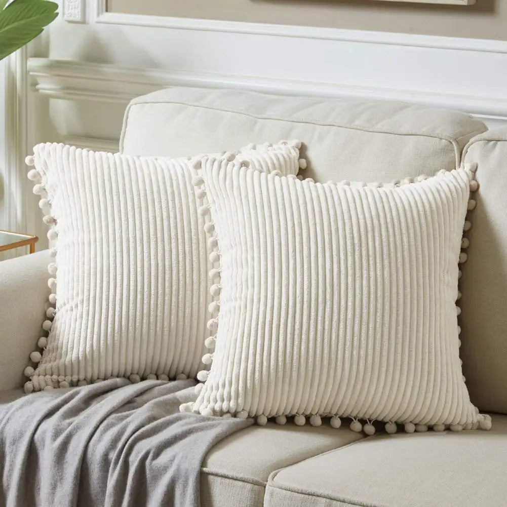 Two white textured pillows with pom-pom details on a beige sofa.