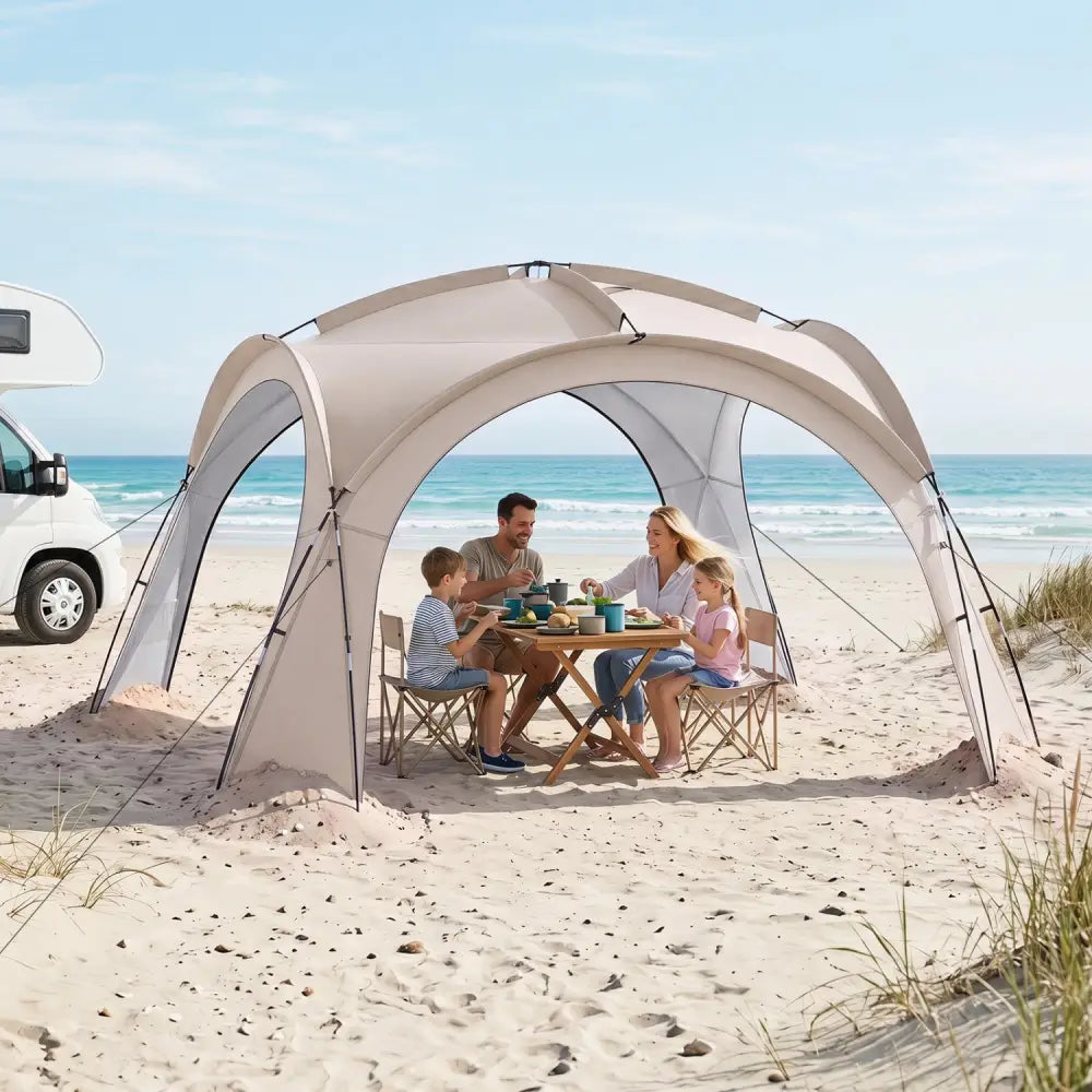 A beige beach canopy tent is set up on a sandy beach with a family dining underneath on folding chairs, with the ocean and sky in the background.