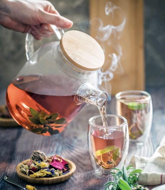 Tea being poured from a glass teapot into a cup with steam rising, surrounded by tea leaves and a wooden board.